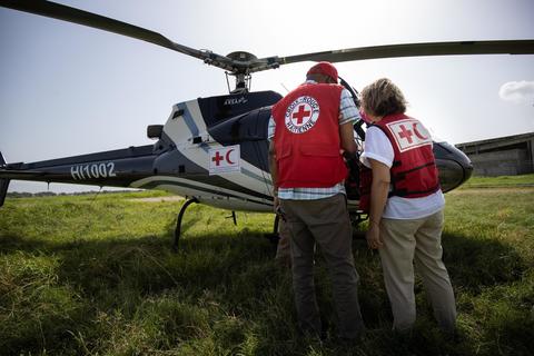 Two people standing in front of a helicopter with their back to the camera, both wear Red Cross / Red Crescent societies gear
